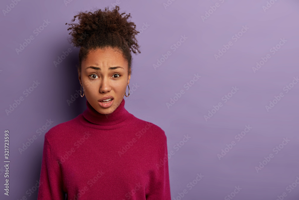 Human facial expressions concept. Displeased Afro woman smirks face and purses lips, reacts on hearing something unpleasant, wears casual jumper, models over purple wall with blank space aside - obrazy, fototapety, plakaty 