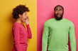 Emotive young curly woman stands in profile and screams loudly at guilty man who has embarrassed face expression, made big mistake, wear bright jumpers, isolated on pink and yellow stuio wall