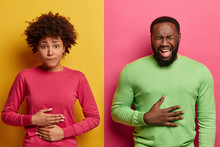Horizontal Shot Of Dark Skinned Couple Touch Stomachs, Suffer From Chronic Gastritis, Being Hungry Has Displeased Face Expressions, Isolated Over Yellow And Pink Background. Healthcare Concept