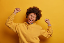 Relaxed Lazy Upbeat Woman Keeps Arms Raised Up In Air, Enjoys Weekends, Dances Joyfully On Disco Party, Closes Eyes And Squints Face, Wears Hoodie, Feels Alive, Poses Over Vibrant Yellow Background