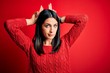 © Krakenimages.com - Young brunette woman with blue eyes wearing casual sweater over isolated red background Doing bunny ears gesture with hands palms looking cynical and skeptical. Easter rabbit concept.