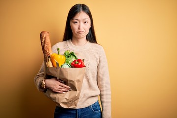 Young asian woman holding paper bag of fresh healthy groceries over yellow isolated background depressed and worry for distress, crying angry and afraid. Sad expression.