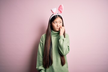 Sticker - Young asian woman wearing cute easter bunny ears over pink background looking stressed and nervous with hands on mouth biting nails. Anxiety problem.