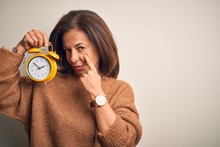 Middle Age Brunette Woman Holding Clasic Alarm Clock Over Isolated Background Pointing To The Eye Watching You Gesture, Suspicious Expression
