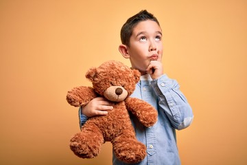 Wall Mural - Young little boy kid hugging teddy bear stuffed animal over yellow background serious face thinking about question, very confused idea