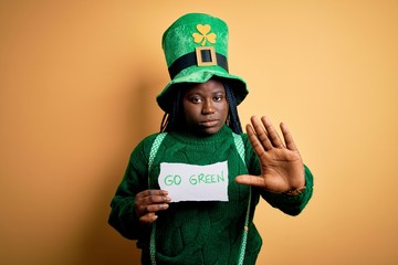 Poster - Plus size african american woman wearing green hat holding paper on saint patricks day with open hand doing stop sign with serious and confident expression, defense gesture