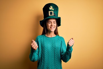 Young beautiful brunette woman wearing green hat with clover celebrating saint patricks day very happy and excited doing winner gesture with arms raised, smiling and screaming for success. Celebration