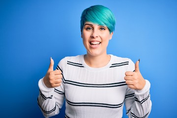 Young beautiful woman with blue fashion hair wearing casual sweater standing at studio success sign doing positive gesture with hand, thumbs up smiling and happy. Cheerful expression and winner