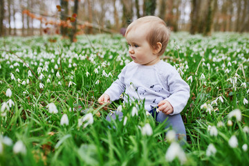 Wall Mural - Cute one year old baby girl sitting on the grass with many snowdrop flowers in park or forest