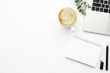 White minimalist office desk table with blank notebook page with pen, cup of latte coffee and laptop computer. Top view with copy space, flat lay.
