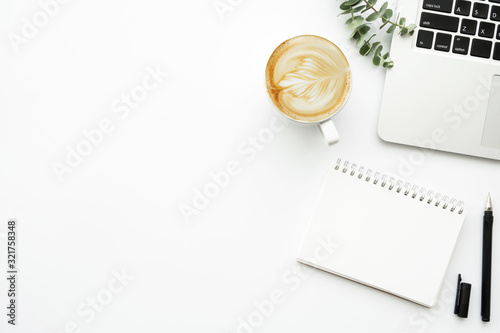 White minimalist office desk table with blank notebook page with pen, cup of latte coffee and laptop computer. Top view with copy space, flat lay.