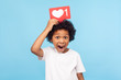 © khosrork - Cute surprised funny little boy with curly hair holding heart Like icon on his head and open mouth in amazement shock, showing button of social network. indoor studio shot isolated on blue background