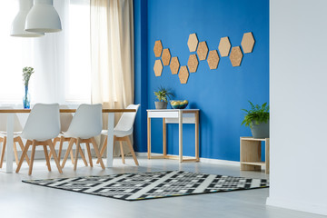 Wall Mural - Cork honeycombs on blue wall of trendsetting dining room interior with patterned carpet, plants and white furniture