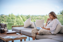 Attractive Woman 40 Years Old In A White Shirt Sitting On A Gray Sofa Working On A Laptop On The Terrace Overlooking The Green Jungle On A Bright Sunny Day