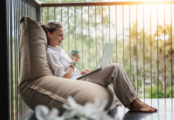 Happy beautiful young woman in a white shirt sits an  bean bag chair working on a laptop on the terrace overlooking the green jungle on a bright sunny day