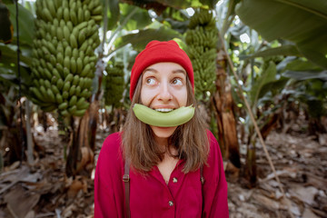 Wall Mural - Portrait of a cute woman dressed in red biting banana fruit on the plantation. Fresh fruits eating concept