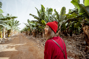 Canvas Print - Woman as a tourist or farmer dressed casually in red shirt and hat walking on the banana plantation with a rich harvest. Concept of green tourism or exotic fruits growing