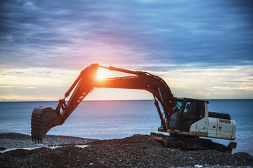 backhoe or digger working with bucket at industrial earth excavation site near sea in sunrise light