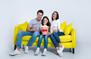 Watching comedies. Happy mother, father and their little cute daughter are sitting together on a yellow couch and laughing while watching comedies.