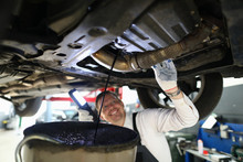 Mechanic Conducts Thorough Inspection Car Garage. Male Mechanic In Working Overall Stands Under Car And Shines Lantern On Car Parts In Search Malfunction. Technician Smiles While Draining Old Oil.