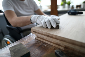 Man sits in a wheelchair and works as carpenter. Small business for disabled people training study concept. Male hand hold emery sponge and polishes wood closeup