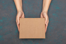 Female Hands Holding Cardboard Box With Empty Copy Space Topsheet Over Dark Wooden Background.