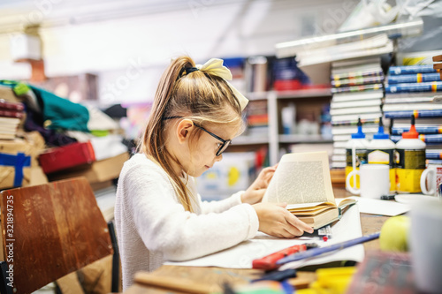 Photographie Adorable caucasian blond girl with ponytail and with eyeglasses sitting in bookstore and reading exciting book