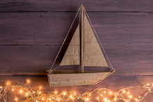 Travel And Adventure Creative Concept - Toy Boat On A Wooden Background. Christmas Lights As A Sea Waves