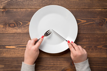 Female hands with knife and fork over white empty plate. Wooden table