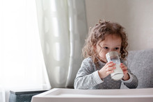 Curly Child Dressed Pajamas At Home Room And Drink Milk At The Morning.. Girl Holding Glass And Liks Tonque Lips. Happy Kid Love Tasty Milk On Bed Table