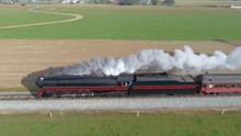 Aerial Side View Of A Restored Steam Engine Blowing Steam And Smoke While Pulling Passenger Cars With View Of Farmlands On A Sunny Day