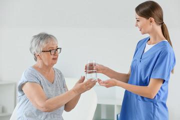 Wall Mural - Female doctor giving senior woman suffering from Parkinson syndrome glass of water in clinic