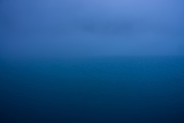 Wall Mural - Natural texture of deep blue calm water in dusk close up. Night sea of blue classic color. Water ripple nature background. Meditative image of thick fog above lake. Soft light shines on water surface.