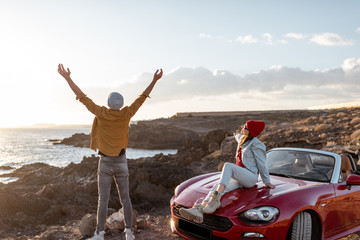 Couple enjoying beautiful views on the ocean, standing together near the car on the rocky coast. Carefree lifestyle, love and travel concept