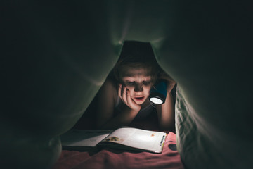 surprised little girl is read a book under a blanket with a flashlight in a dark room at night, in bed, ton