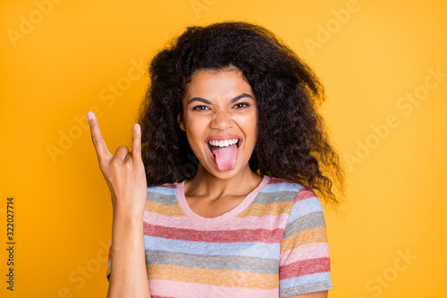 Close-up portrait of nice attractive funky cheerful cheery crazy wavy-haired girl in striped t-shirt showing tongue out horn sign symbol isolated on bright vivid shine vibrant yellow color background