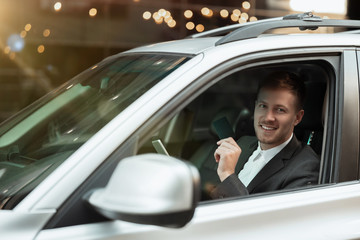 Wall Mural - young smiling businessman looks happy while driving his car with open window, multitasking, big city life