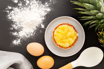 Wall Mural - Pineapple tart or tartlet with grated coconut by an entire pineapple fruit, spilled flour, grey napkin, eggs and cooking spoon, all on a slate background. Flat lay.