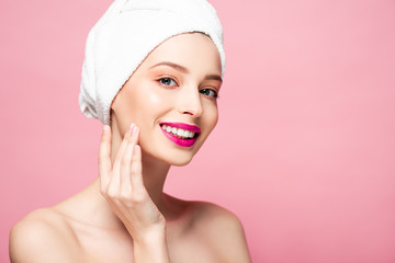 smiling naked woman in white towel touching face isolated on pink