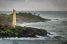 Nawiliwili, Kauai, Hawaii, USA. - January 16, 2020: Yellow Ninini Lighthouse On Black Ocean-shore Rocks Early Morning. Dark Gray Ocean Under Light Gray Sky. Some Green Foliage On Land.