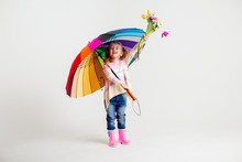 Little Blonde Girl Is Smiling, Standing In Rubber Boots Holding Spring Flowers And A Multi-colored Umbrella On A White Background