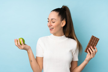 Wall Mural - Young brunette woman over isolated blue background taking a chocolate tablet in one hand and an apple in the other