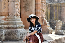 Young Pretty Asian Girl Dressed In A Striped Dress And White Hat Sits On The Foundation Of An Ancient Hindu Temple. Prasat Hin Phanom Rung, Thailand