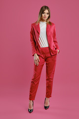 Blonde model in red pantsuit, white blouse, high black heels. Posing standing against pink studio background. Beauty, fashion. Full length