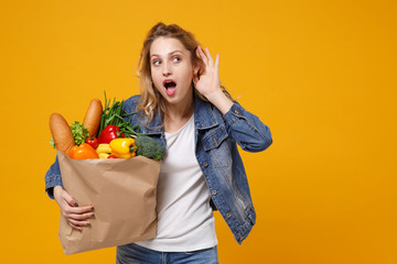 Wall Mural - Shocked young woman in denim clothes isolated on orange background. Delivery service from shop or restaurant concept Hold brown craft paper bag for takeaway mock up with food products try to hear you.