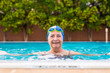 © luciano - Senior woman enjoying practices activity outdoor in the swimming pool.  Blue water and green plants