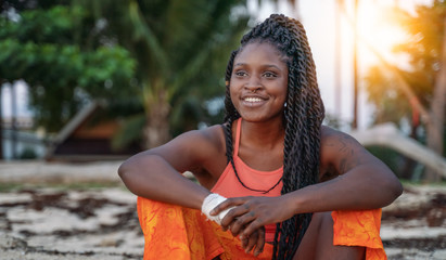 Charming beautiful young african american woman girl with black pigtails with piercings on her face, sitting on the beach during sunset