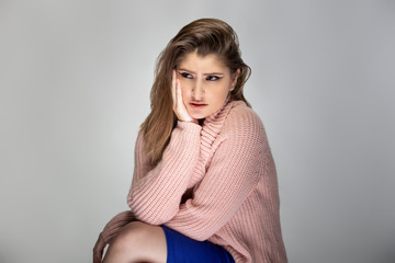 Close up portrait of a young Caucasian woman wearing a pink sweater on a grey background.  She looks sad and depressed