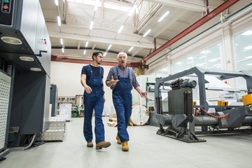 Wall Mural - Experienced senior man in blue overall gesturing hands while interacting with young worker at printing factory