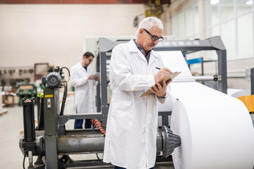 Serious senior inspector in lab coat standing at printing press with roll paper and making notes in clipboard